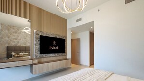 A modern hotel room with a large bed, a mounted TV, a wooden panel wall, and a breakfast tray with fruits and drinks. - Livbnb - 3BR + 1 with Burj and Fountain Views (Dubai)