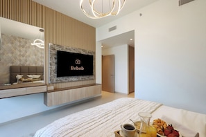 A modern hotel room with a large bed, a mounted TV, a wooden panel wall, and a breakfast tray with fruits and drinks.