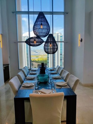 Uma sala de jantar com uma mesa comprida posta para seis pessoas, grandes janelas e luminárias pendentes.