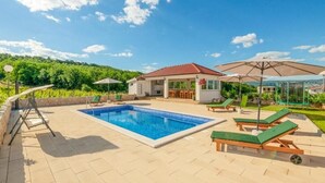 Une piscine entourée de chaises longues et de parasols, elle-même bordée d’un jardin aménagé et d’un petit bâtiment doté d’un toit rouge.