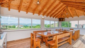 Un vaste espace dédié à la salle à manger, avec une table et des bancs en bois, de grandes fenêtres offrant une vue sur la campagne et un plafond en bois.