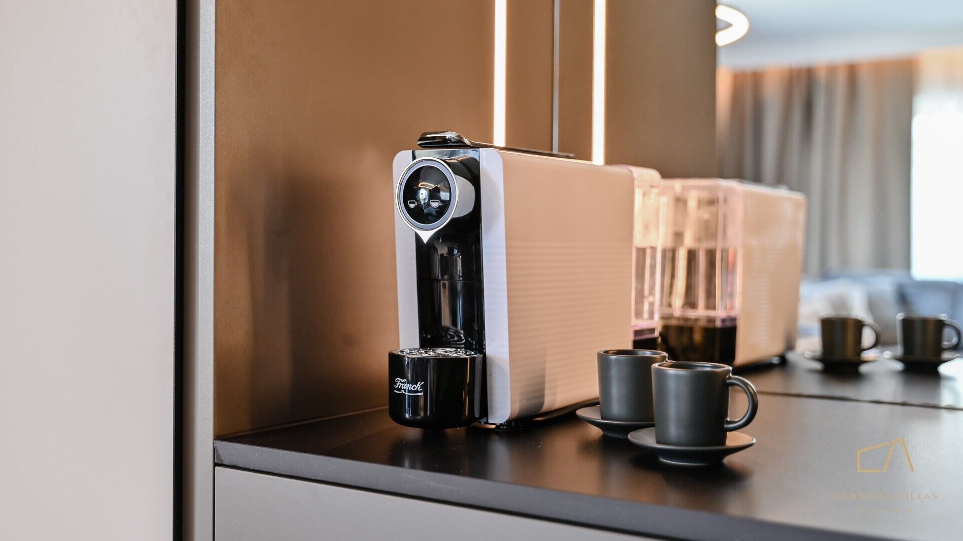 Een moderne koffiemachine met een digitaal scherm, twee kopjes en een glas koffie op een aanrecht.