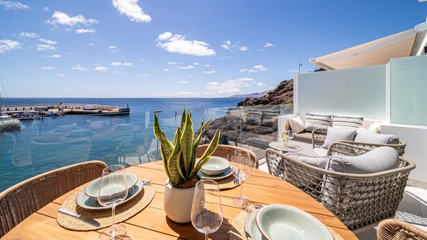 A dining table with plates, glasses, and a potted plant, overlooking a coastal view with boats and a rocky shoreline.