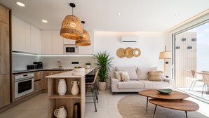 A modern kitchen with white cabinets, a dining area with a white sofa and a coffee table, and a large window providing natural light.