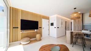 A modern living room with a flat-screen TV, wooden panel wall, round dining table, and a kitchen area with bar stools.