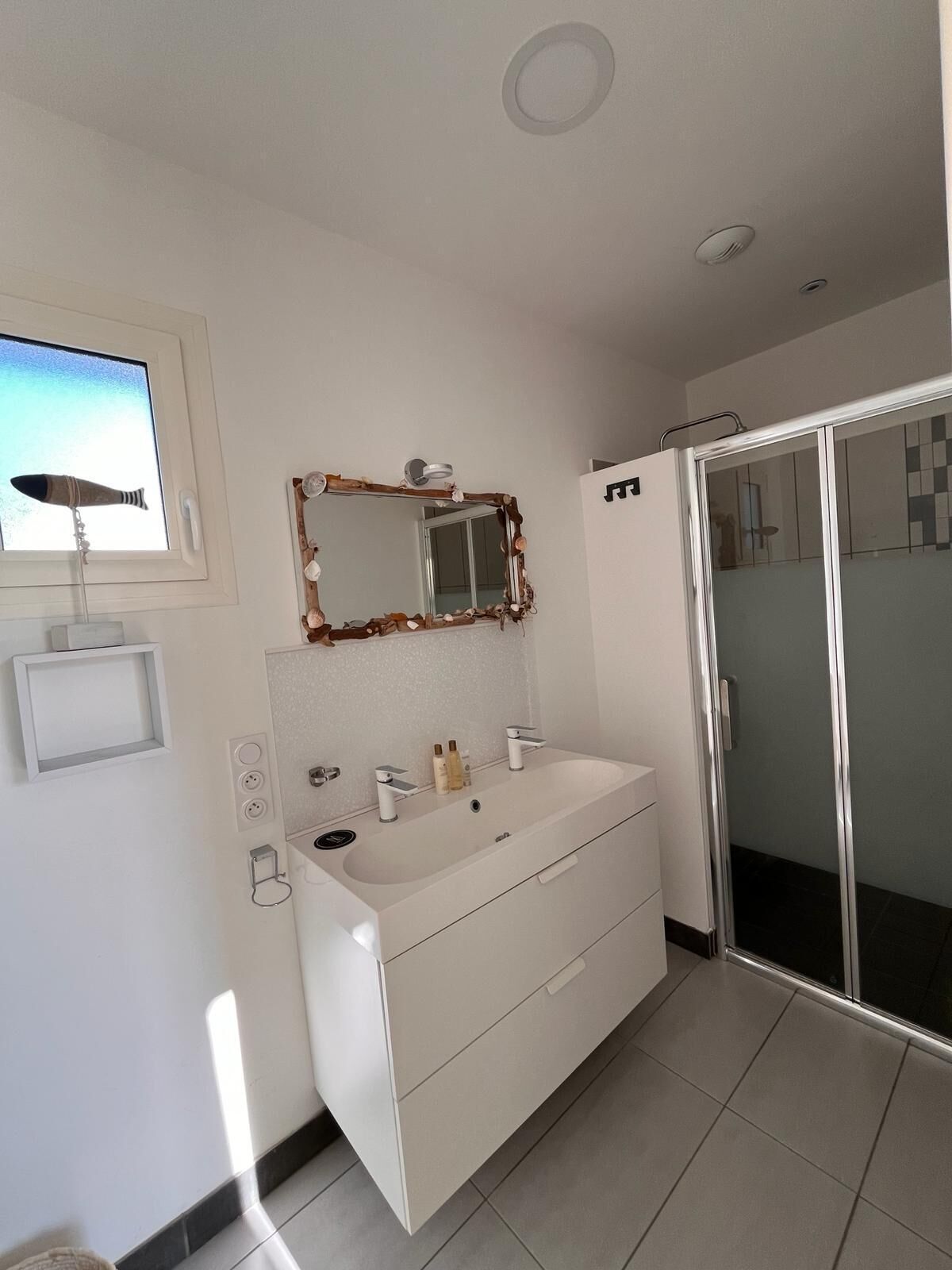Une salle de bain moderne avec un meuble-lavabo blanc, un grand miroir, une douche avec une porte en verre et une fenêtre donnant sur le ciel.