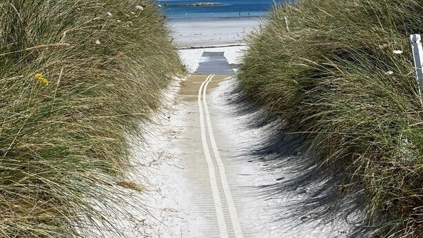 Un sentier de sable qui longe la plage et mène à la mer, bordé d’herbes hautes.