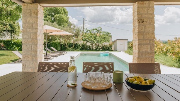 A wooden table with a bottle, glasses, and a bowl of lemons under a pergola with a pool in the background. - Casa Amalia with saltwater pool (Visnjan)