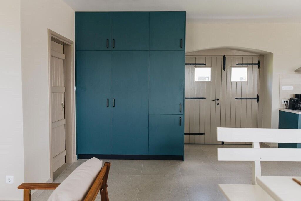 A room with a teal wardrobe, a white bench, and a kitchen area with a window.