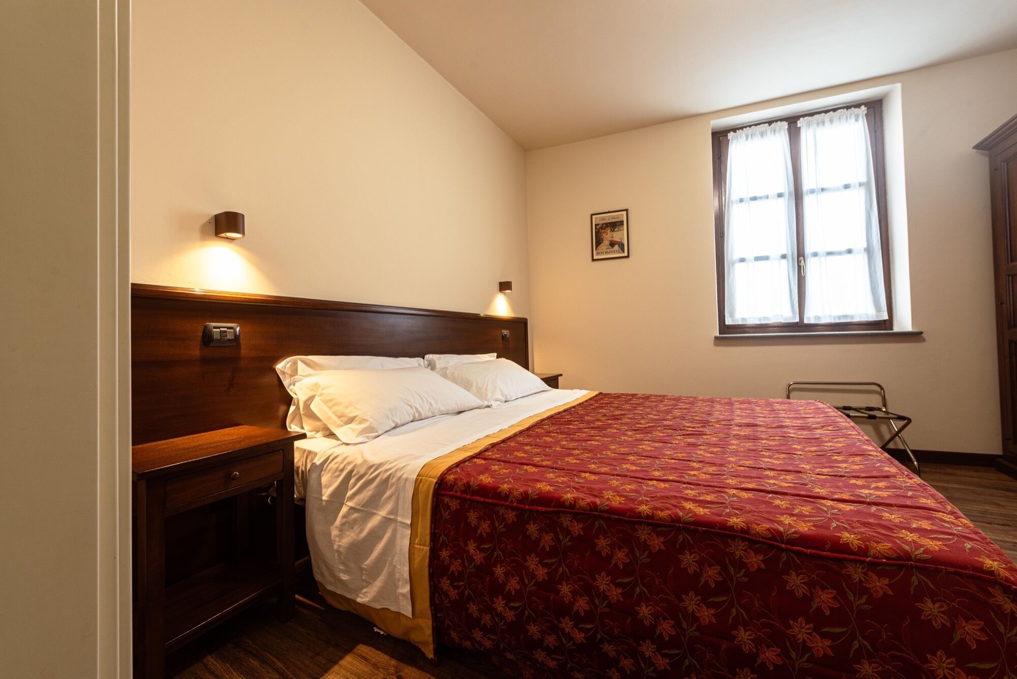 Une chambre à coucher avec une tête de lit en bois, un lit recouvert de draps blancs, une couvre-lit à motifs floraux rouges, une table de chevet, une lampe fixée au mur, une fenêtre avec des rideaux et un petit tableau encadré accroché au mur.