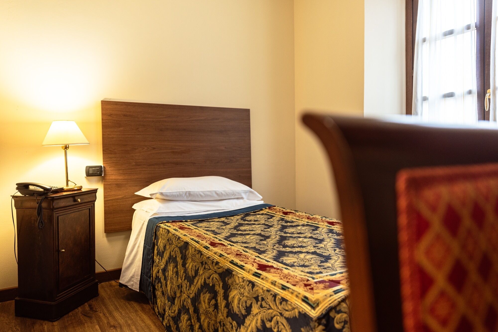 A hotel room with a bed, a nightstand with a lamp, and a wooden headboard.