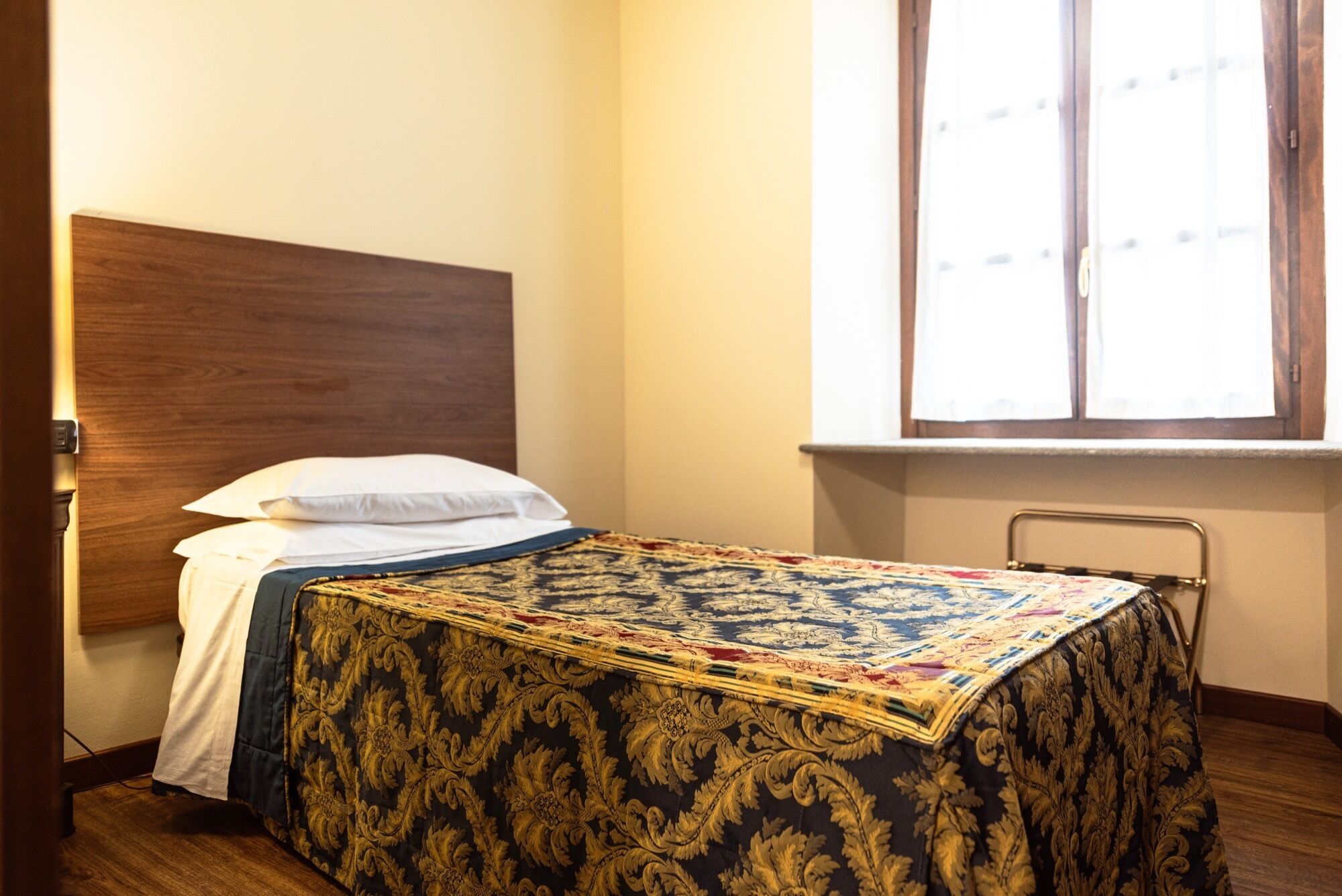 A single bed with a wooden headboard, a white pillow, and a patterned bedspread. There is a window with blinds to the right of the bed.