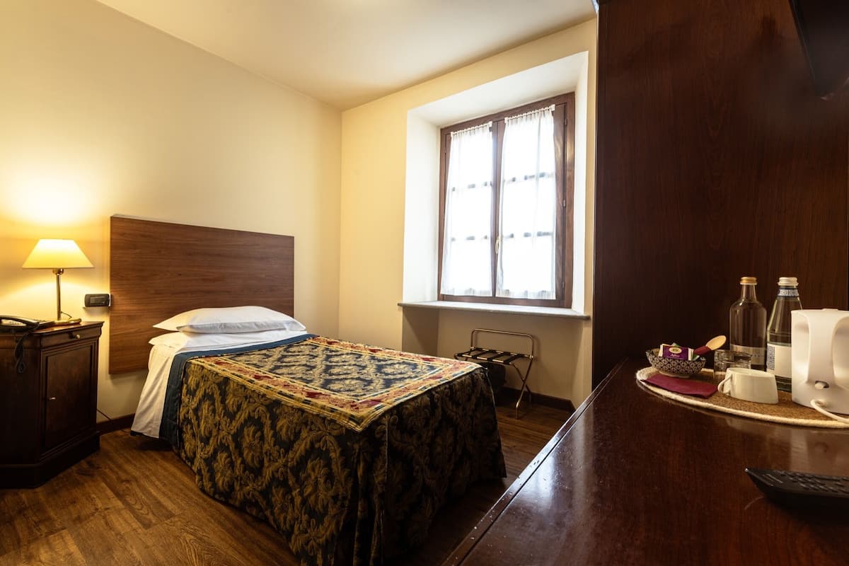 A hotel room with a bed, bedside table, lamp, window, and a small table with a bottle and glasses.