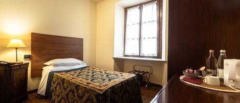 A hotel room with a bed, bedside table, lamp, window, and a small table with a bottle and glasses.