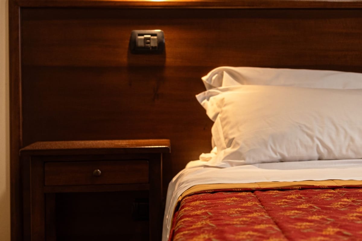 Ett hotellrum med en sänggavel i trä, ett nattduksbord med en lampa och en säng med ett rött, mönstrat överkast.