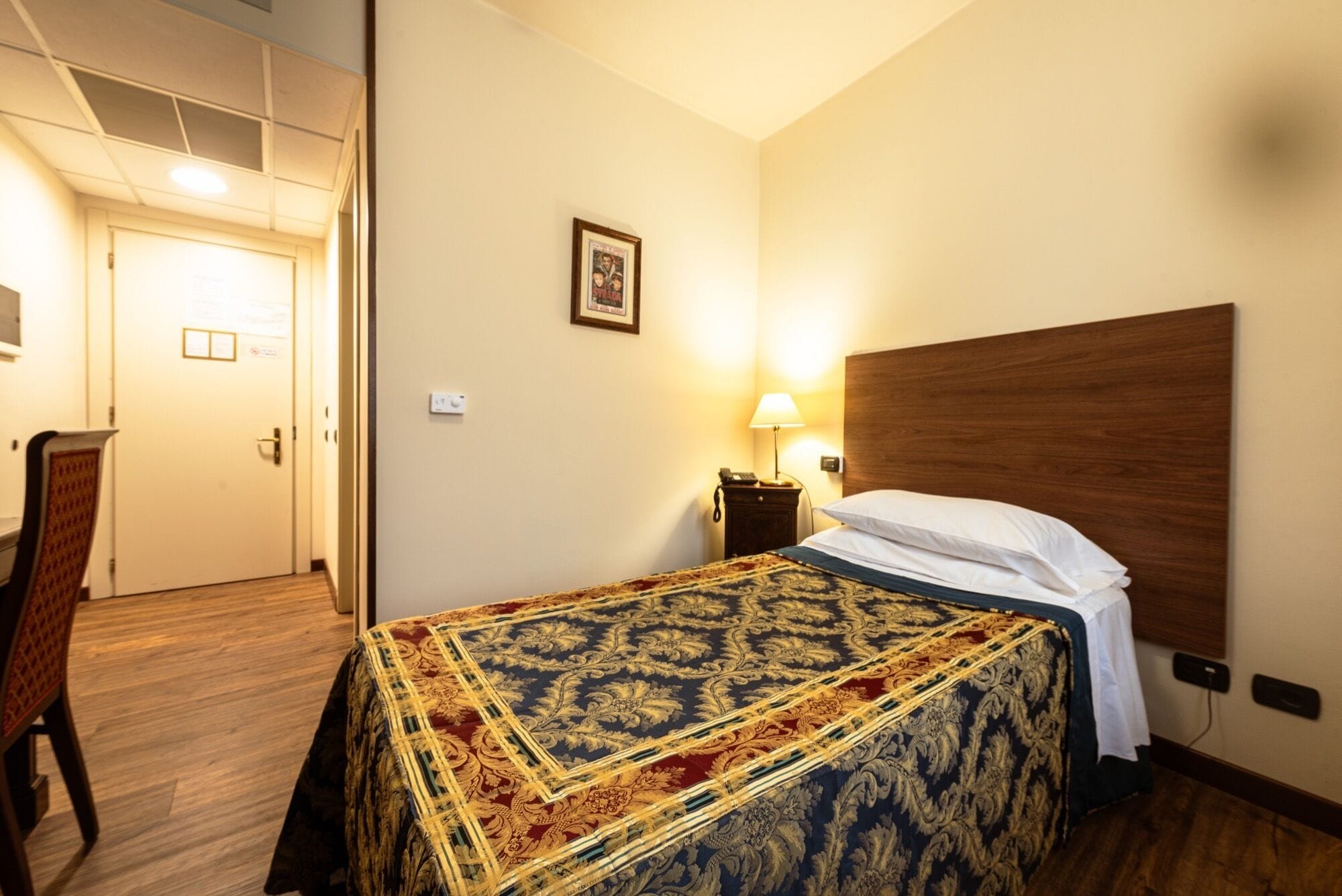 Una habitación de hotel con una cama, una mesita de noche, una lámpara, una silla y una puerta.