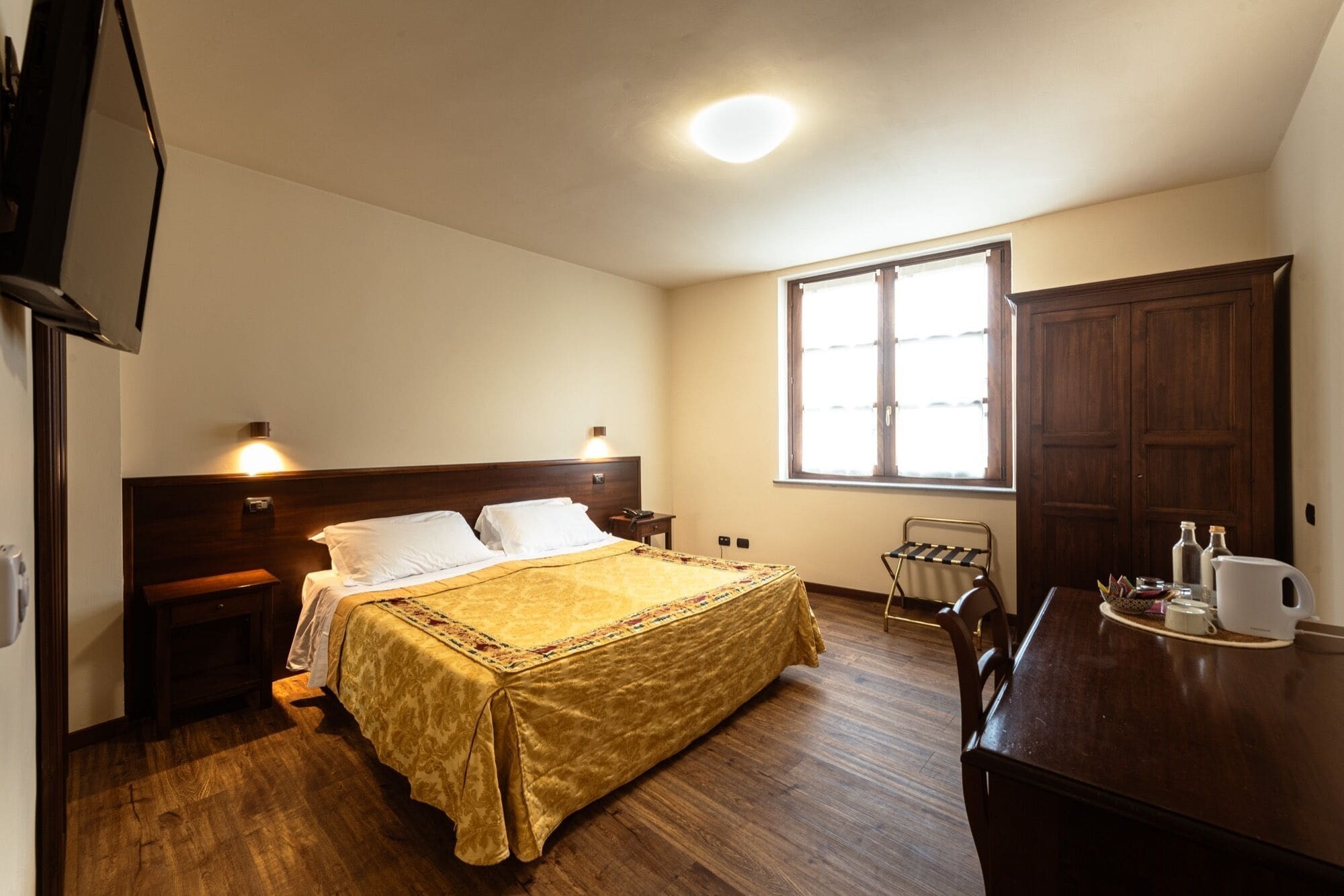 A hotel room with a bed, bedside tables, a desk, a chair, a wardrobe, and a window.