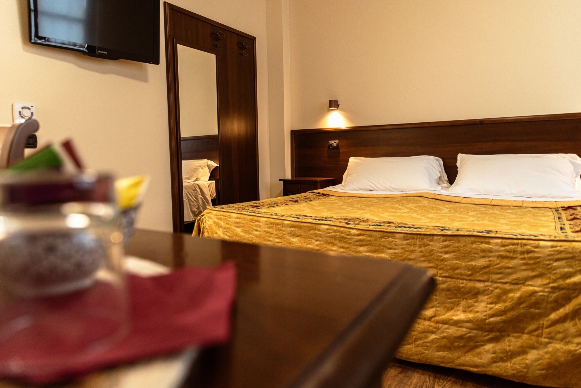 Habitación de hotel con cama, mesita de noche, lámpara, televisor en la pared y espejo.