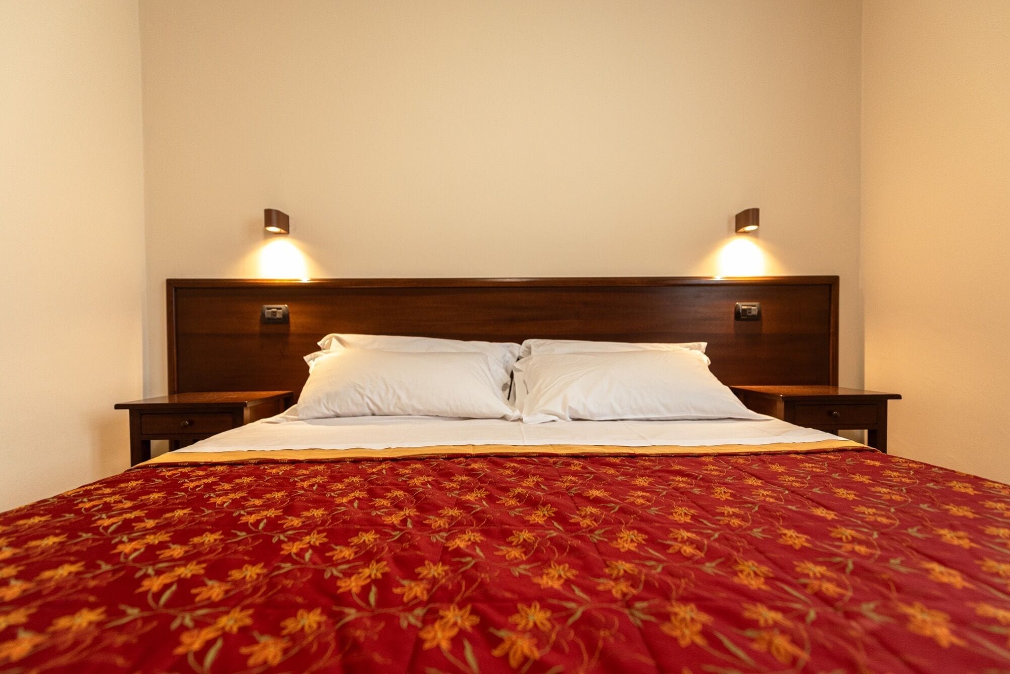 A neatly made bed with a red floral bedspread, two bedside tables, and wall-mounted reading lamps.