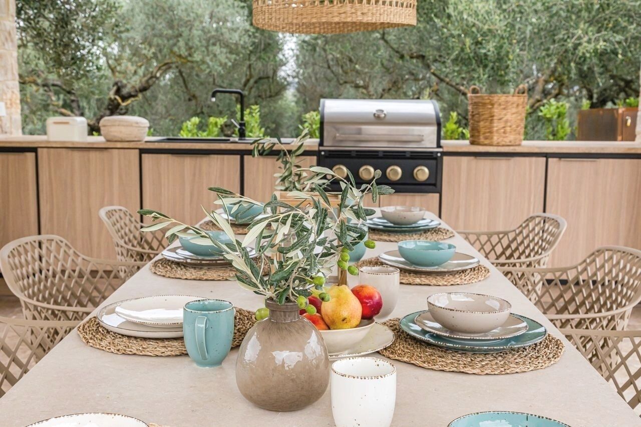 Un espace extérieur aménagé pour les repas, avec une table dressée pour quatre personnes, entourée de chaises en osier. On aperçoit un barbecue à l’arrière-plan, et la table est décorée de plantes en pot et d’un panier en osier.