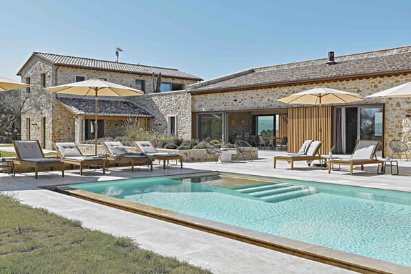A pool area with lounge chairs and umbrellas, stone buildings, and a clear sky.