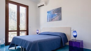 A bedroom with a bed, a nightstand, a lamp, a painting, and a window with a view of mountains. - Casa Orchidea Bianca with large equipped veranda (San Vito Lo Capo)