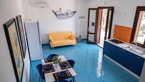 A compact kitchen with blue tiled flooring, a dining area with a table set for two, a yellow sofa, and a small decorative hammock on the wall. - Casa Orchidea Bianca with large equipped veranda (San Vito Lo Capo)
