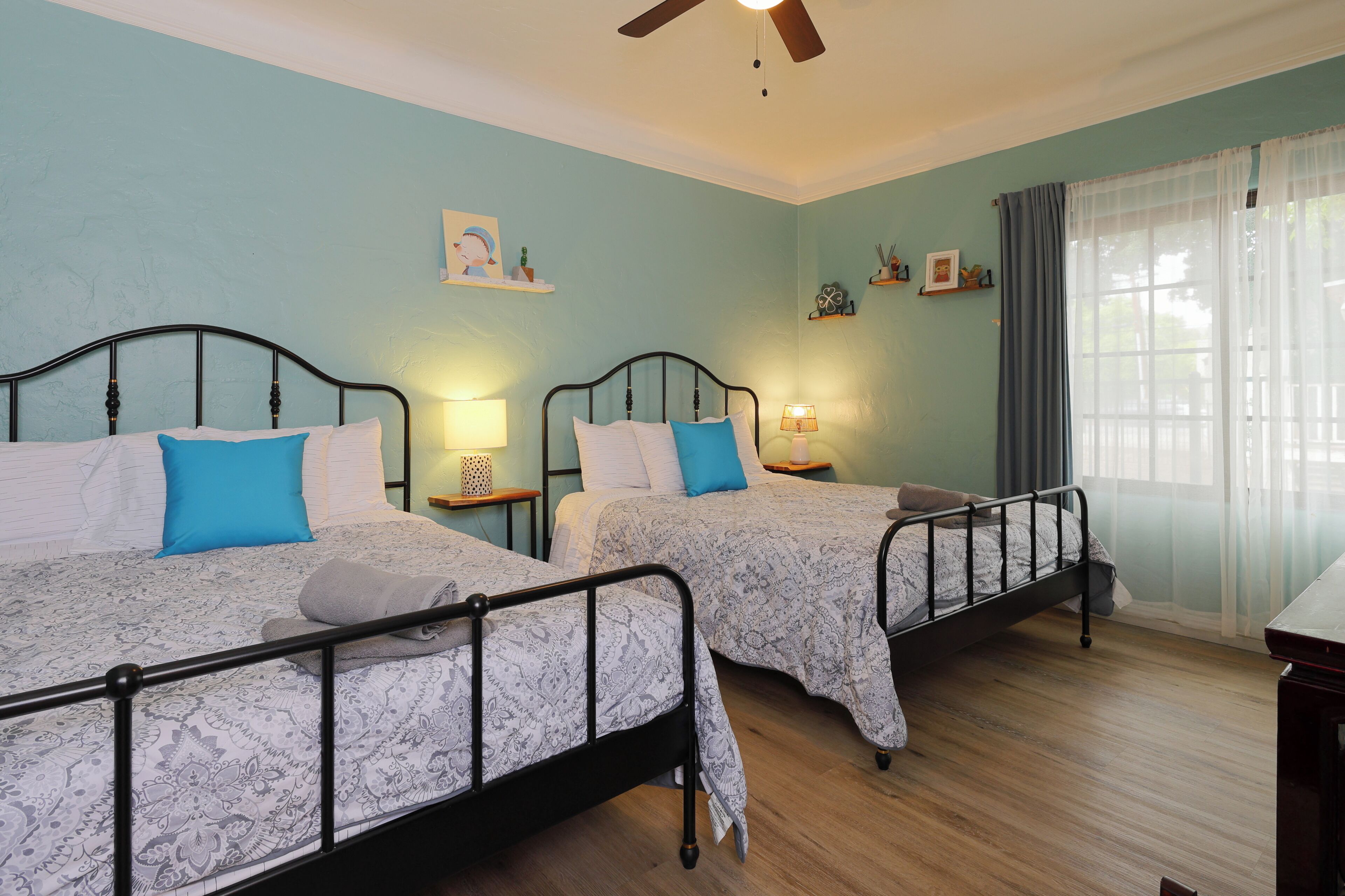 Une chambre à coucher avec deux lits, chacun doté d’une tête de lit, de tables de chevet et de lampes. Les murs de la chambre sont d’un bleu clair, le sol est en bois et il y a une fenêtre avec des rideaux.