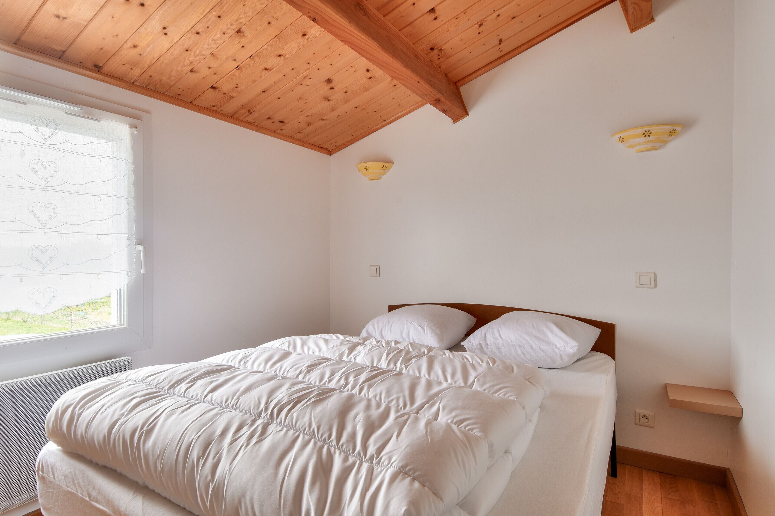 Une chambre avec un plafond en bois, un lit avec une literie blanche, une fenêtre ornée de rideaux à motifs de cœurs et une petite étagère fixée au mur.