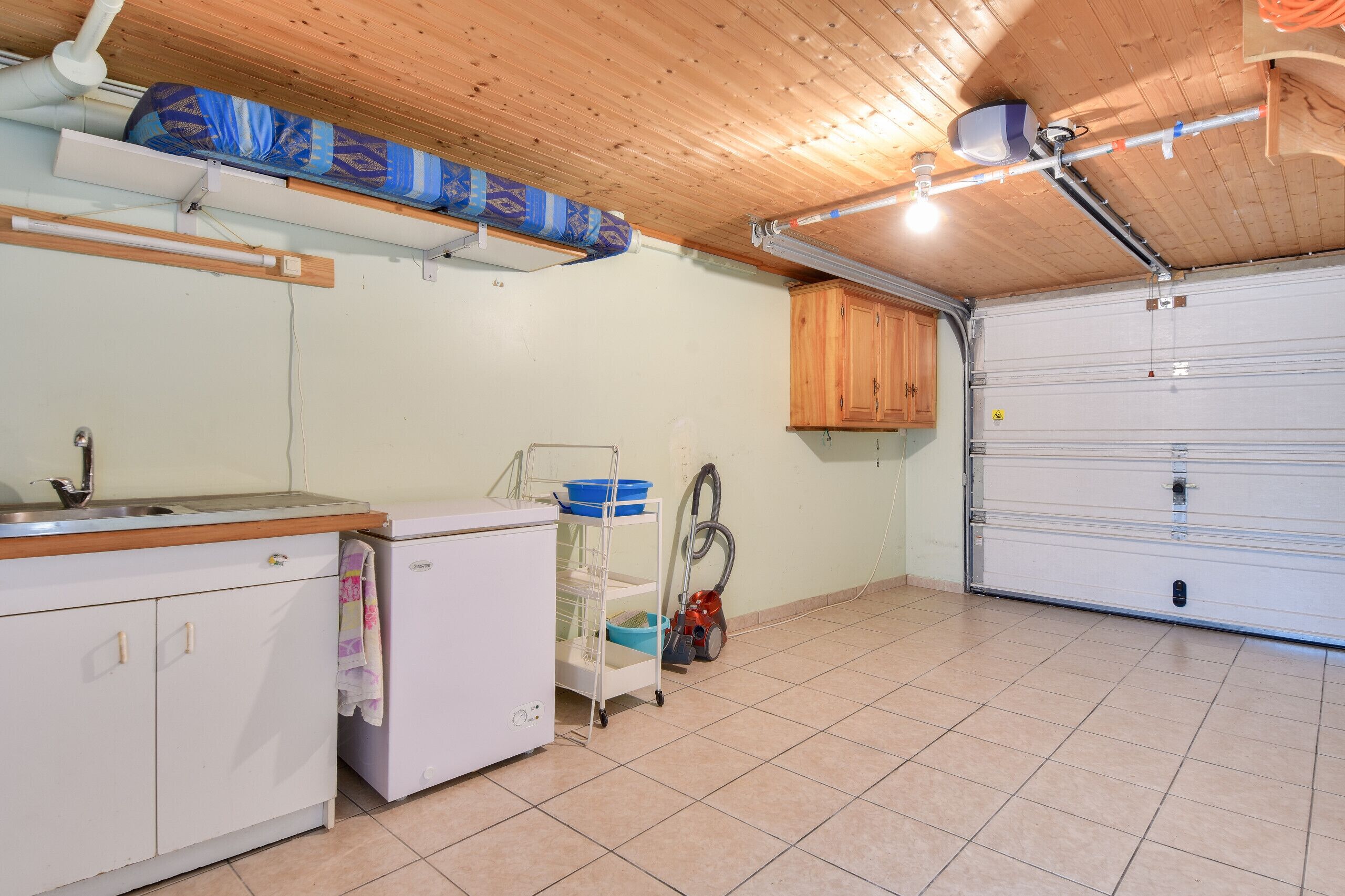 Una cocina con un lavamanos, un refrigerador, un carrito con productos de limpieza y una puerta de garaje.