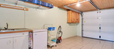 Una cocina con un lavamanos, un refrigerador, un carrito con productos de limpieza y una puerta de garaje.
