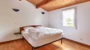 Een slaapkamer met een houten plafond, een bed, een nachtkastje en een raam met uitzicht op groen.