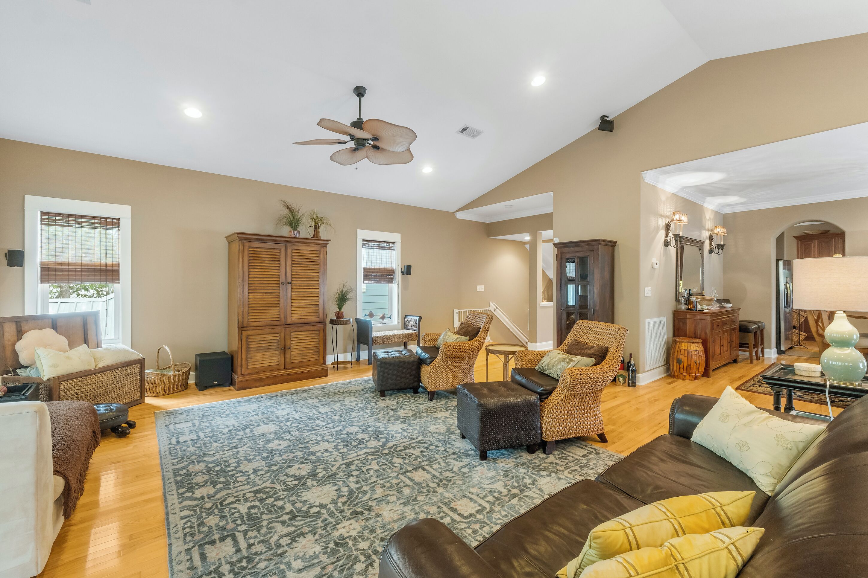 A spacious living room with a high ceiling, a ceiling fan, and a large window. There are multiple seating areas with sofas and chairs, a coffee table, and a dining table.