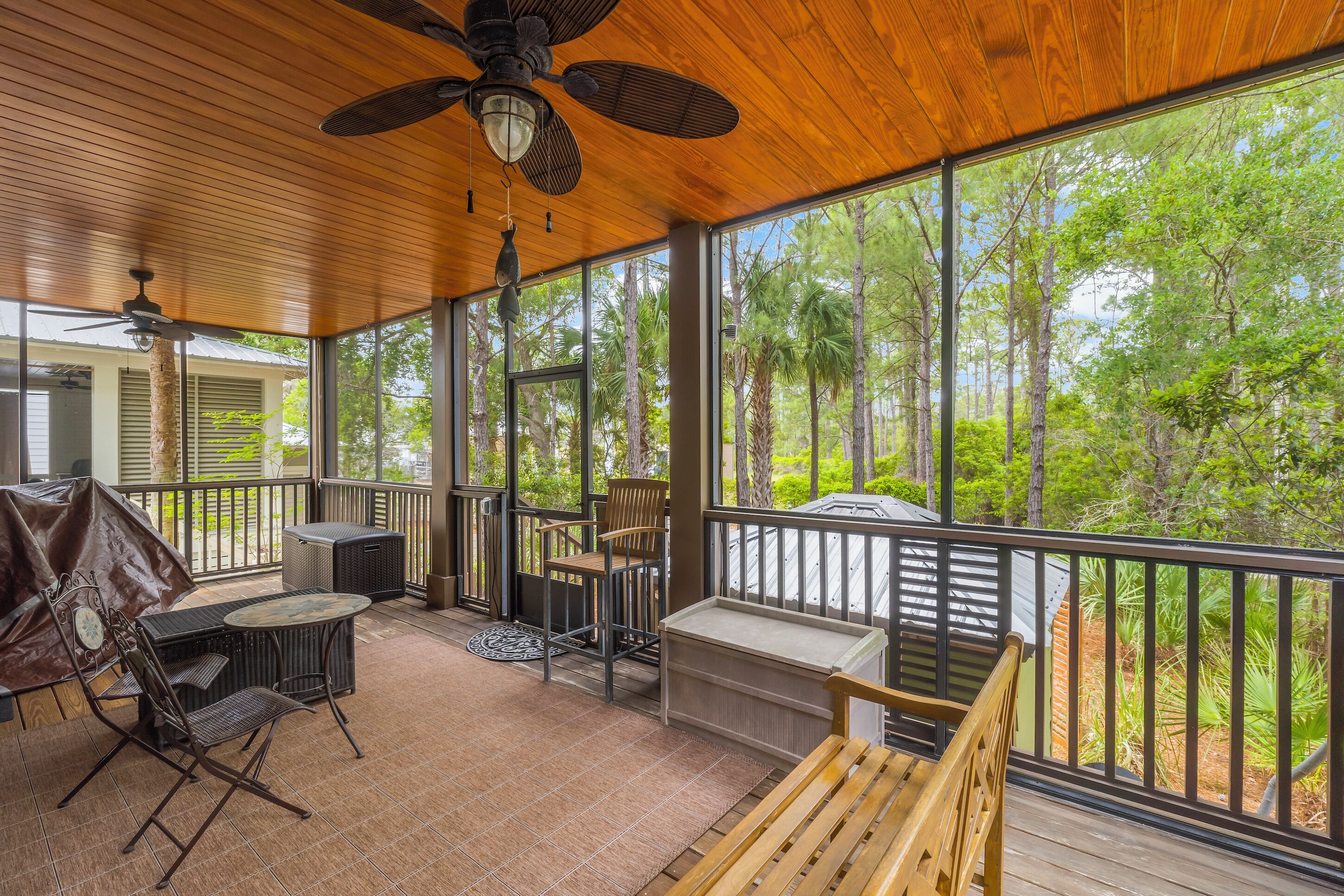 A covered outdoor area with a wooden ceiling, ceiling fans, and large windows offering a view of trees.