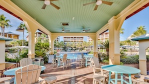 A covered outdoor seating area with tables and chairs, a pool, and a view of hotel buildings in the background.