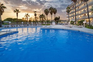 Una piscina con sillones de descanso y palmeras, rodeada por un edificio de hotel con balcones.
