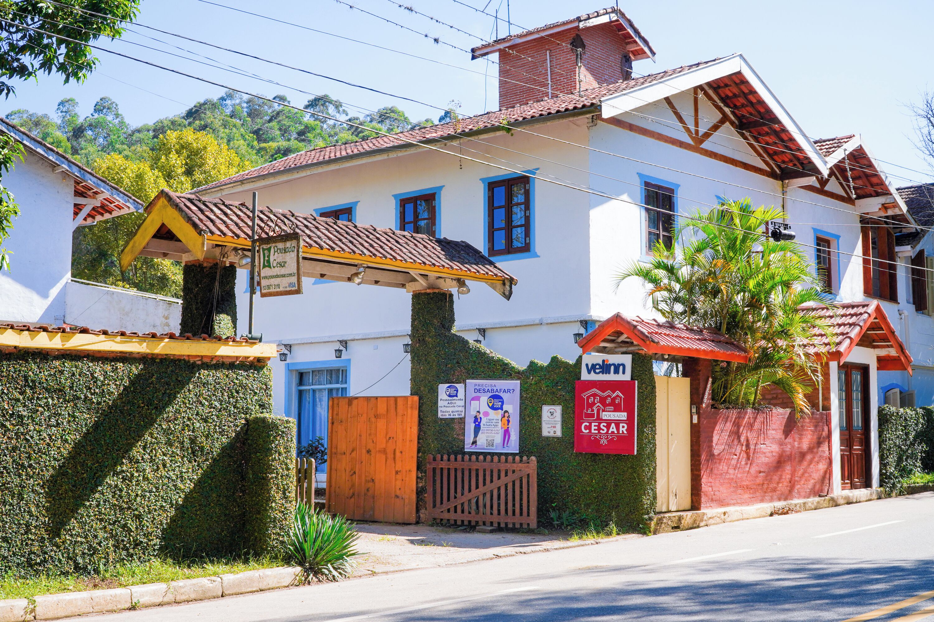 A white building with blue windows and a red roof, surrounded by greenery and a wooden fence.