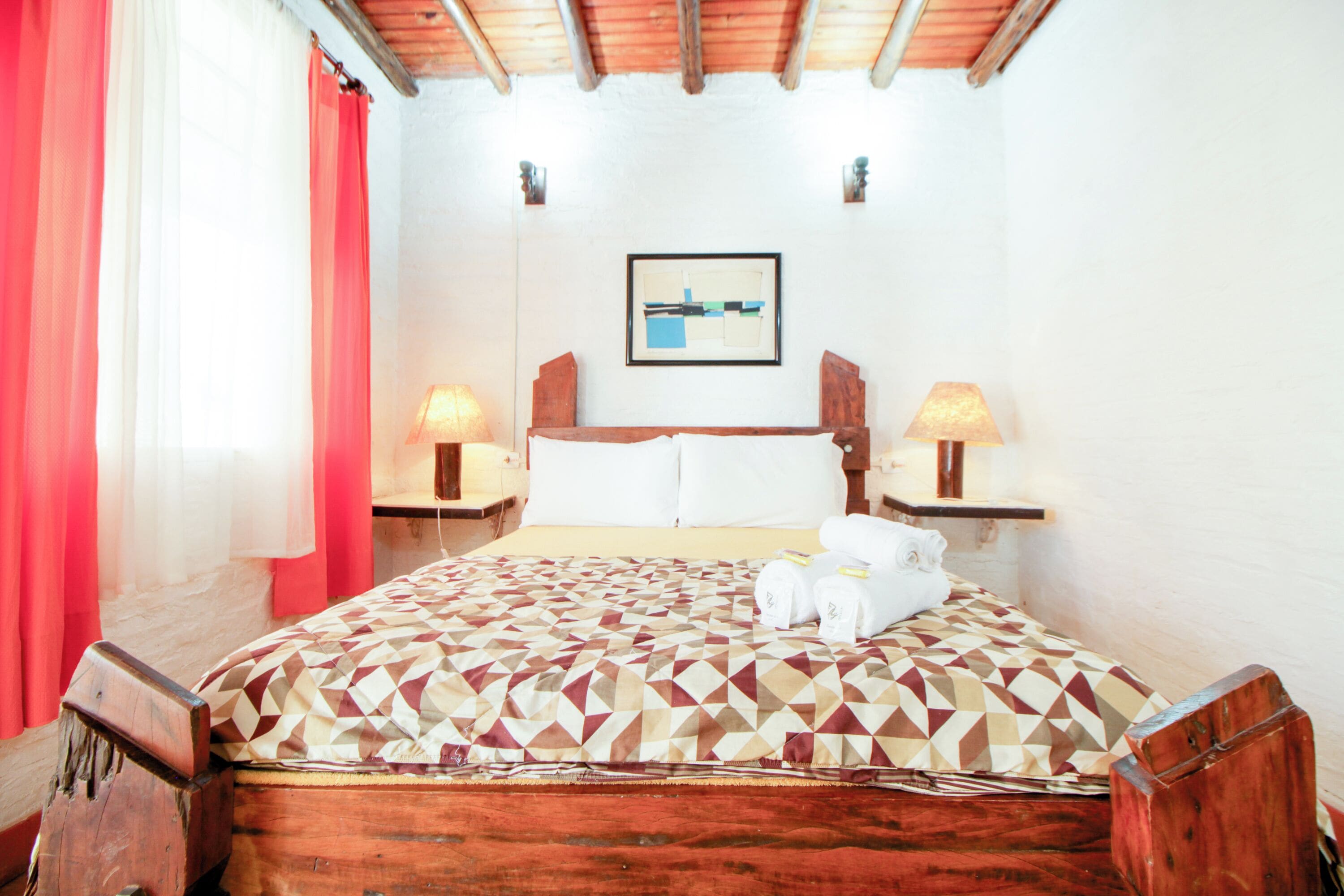 A hotel room with a wooden bed, a patterned bedspread, two bedside lamps, a framed picture on the wall, and a red curtain.