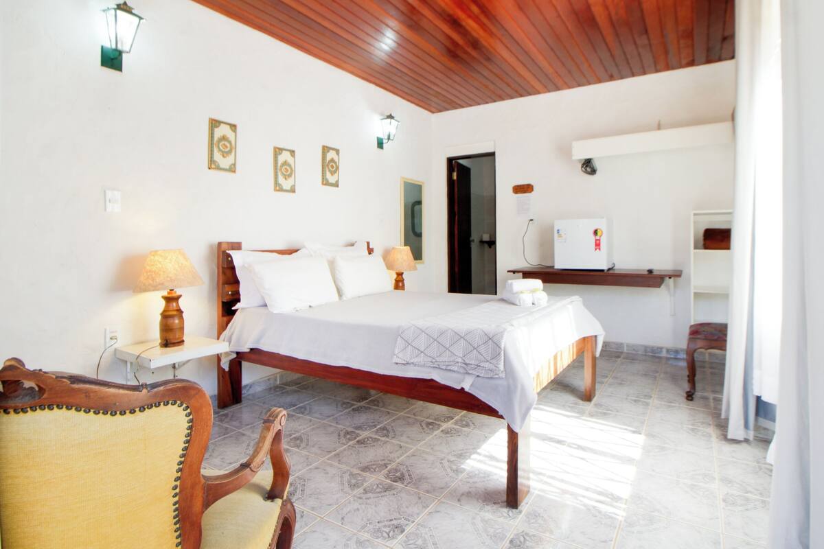 A bedroom with a bed, bedside lamps, a wooden chair, a desk, and a refrigerator.