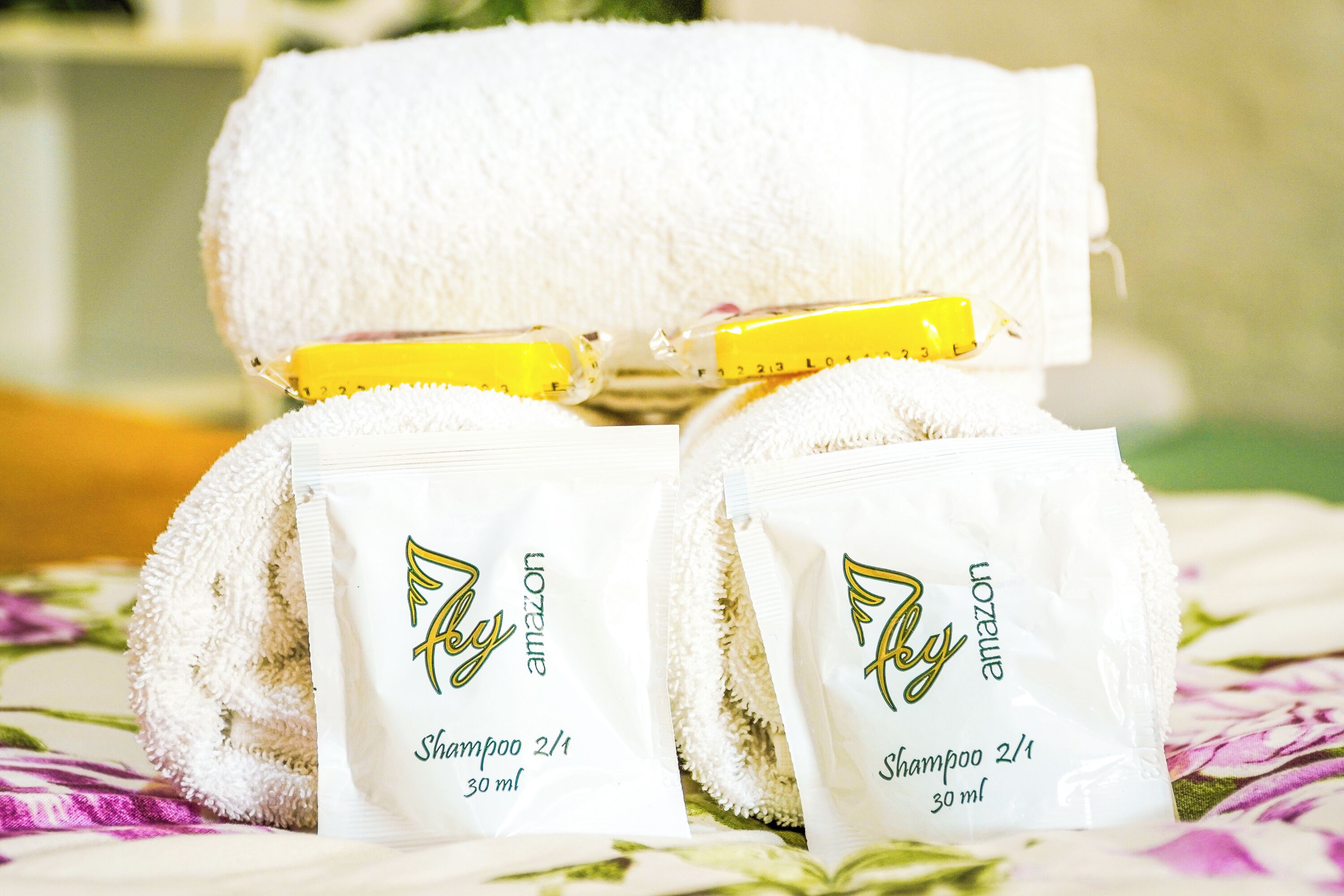 Zwei weiße Handtücher und zwei Shampoo-Beutel mit gelber Kappe auf einem blumengemusterten Bett.