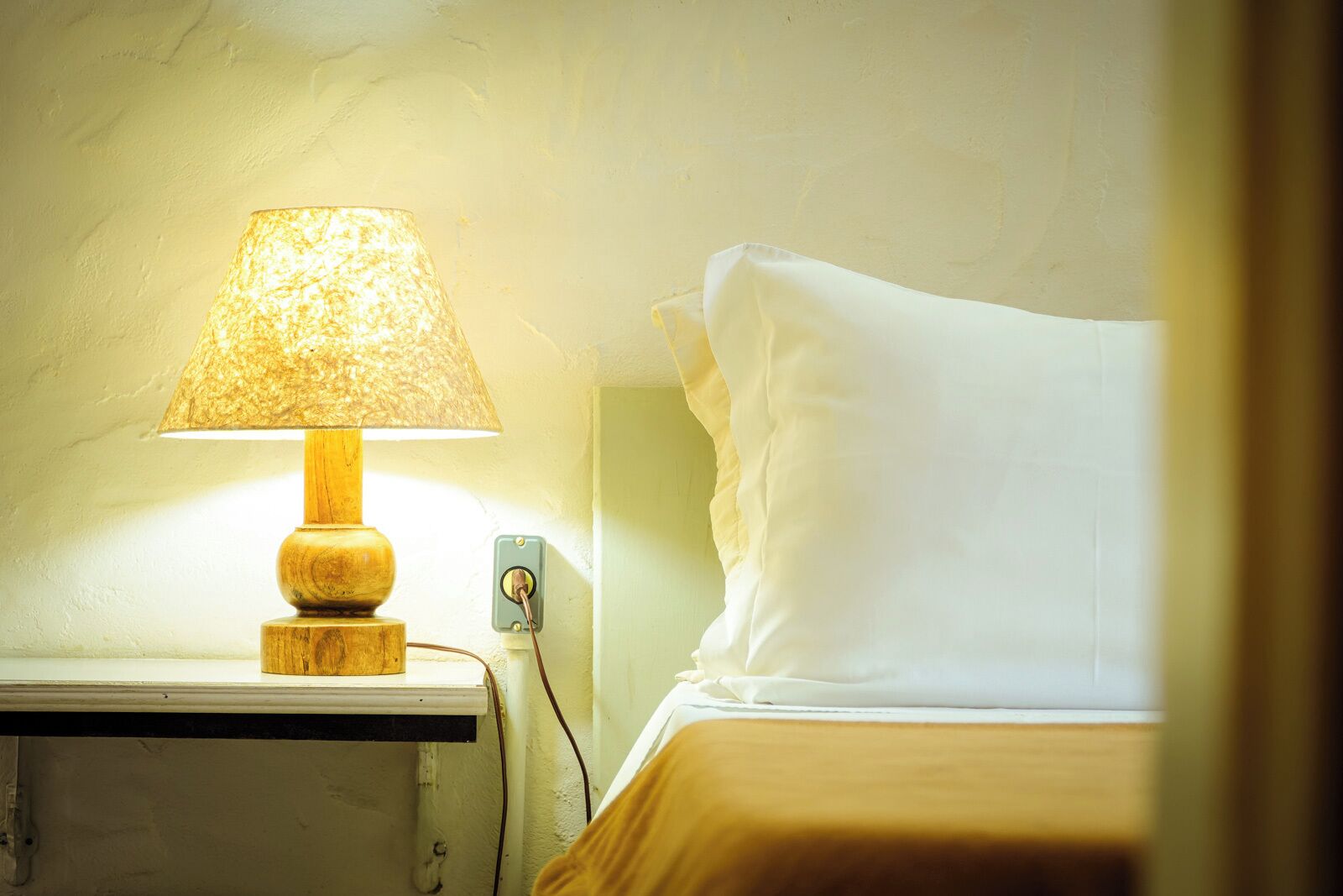 Una cama con un almohadón blanco y una mesita de noche de madera con una lámpara.