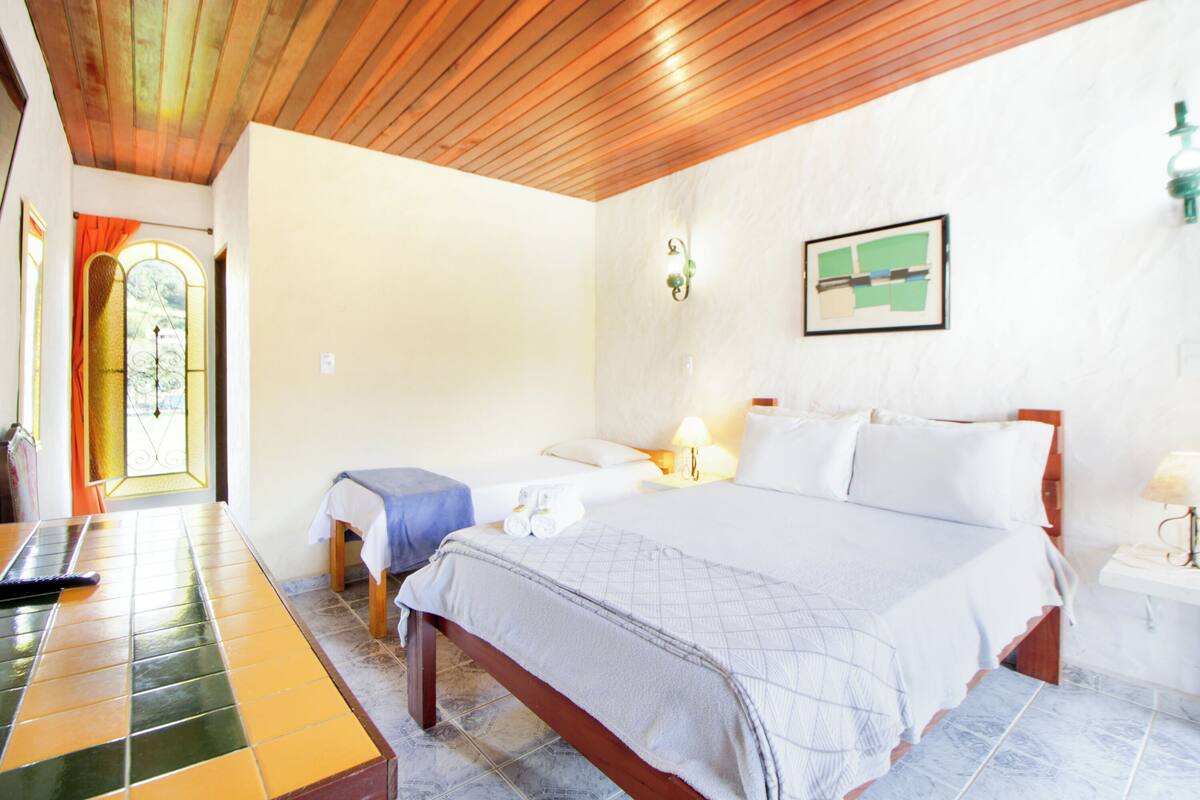 A hotel room with a wooden ceiling, a bed with white linens, a small table with a blue tablecloth, and a window with curtains.