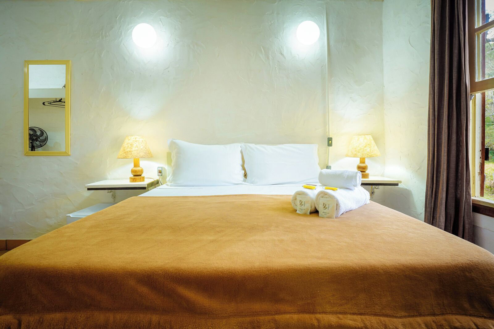 Habitación de hotel con una cama grande, dos lámparas de noche, un espejo y un ventanal con cortinas.