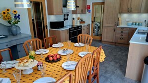 A dining area with a table set for four, surrounded by wooden chairs. There is a kitchen in the background with wooden cabinets and a microwave.