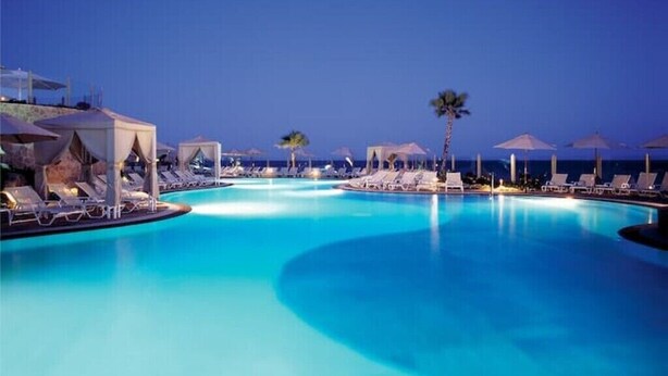 A swimming pool with lounge chairs and umbrellas, surrounded by a low wall and palm trees.