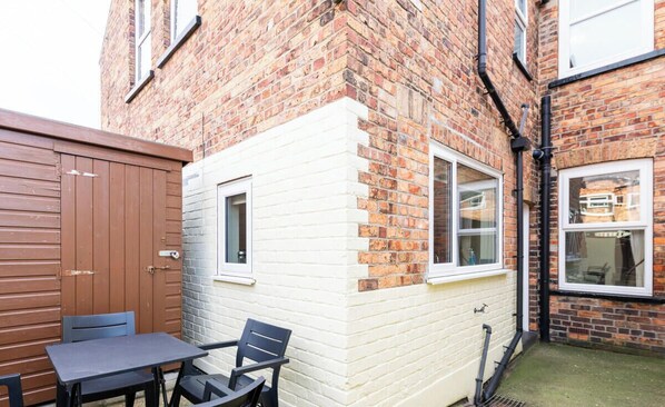 Une maison en briques avec une façade blanche, une porte en bois et un petit patio comprenant une table et des chaises.