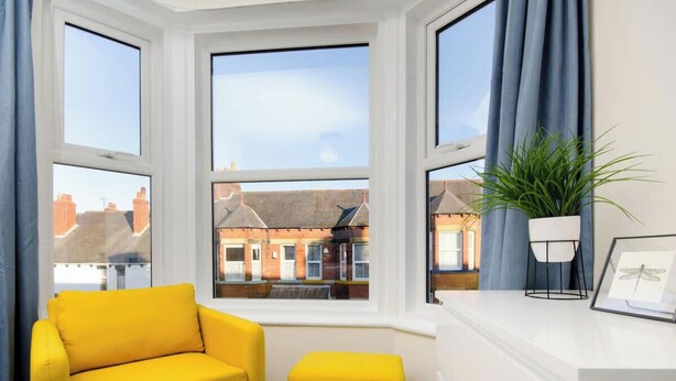 Ein Zimmer mit Erkerfenster, einem gelben Sessel, einer Pflanze und einem Bilderrahmen auf der Fensterbank.