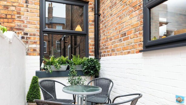 En liten sittplats utomhus med flätade stolar och ett bord i glas, omgiven av planterade växter och en mur av tegelsten.