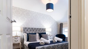 Une chambre à coucher avec une tête de lit capitonnée, un lit avec des serviettes roulées, deux tables de chevet avec des lampes et une fenêtre avec des rideaux.