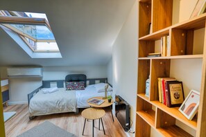 Une chambre à coucher de taille réduite, dotée d’une lucarne, d’un lit, d’une petite table ronde et d’une bibliothèque.